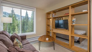 Ein gemütliches Wohnzimmer mit braunem Sofa, einem kleinen Holztisch, einem Fernseher auf einem Holzschrank und einem Bücherregal.