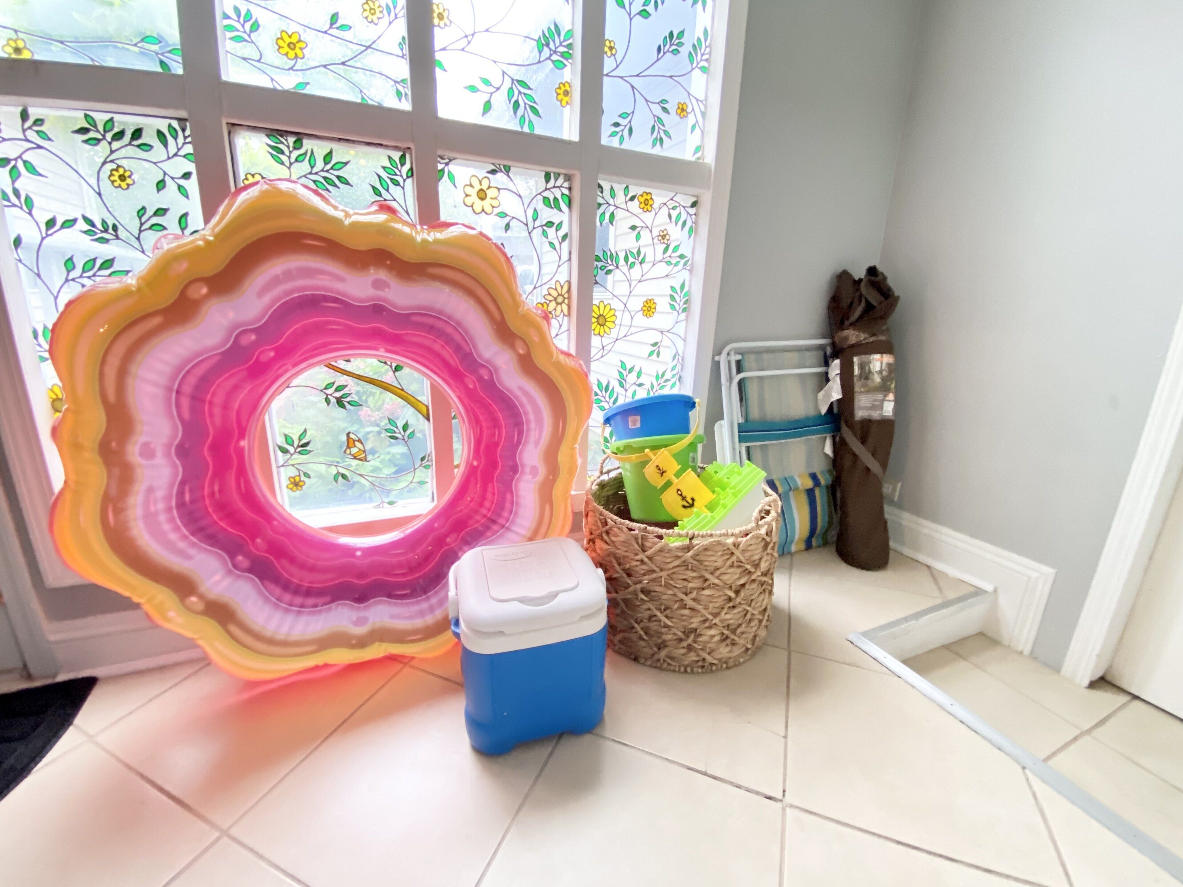Una habitación con una gran pieza de arte de vidrio de colores, una cesta de mimbre, un enfriador azul y una ventana con persianas florales.