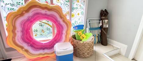Una habitación con una gran pieza de arte de vidrio de colores, una cesta de mimbre, un enfriador azul y una ventana con persianas florales.