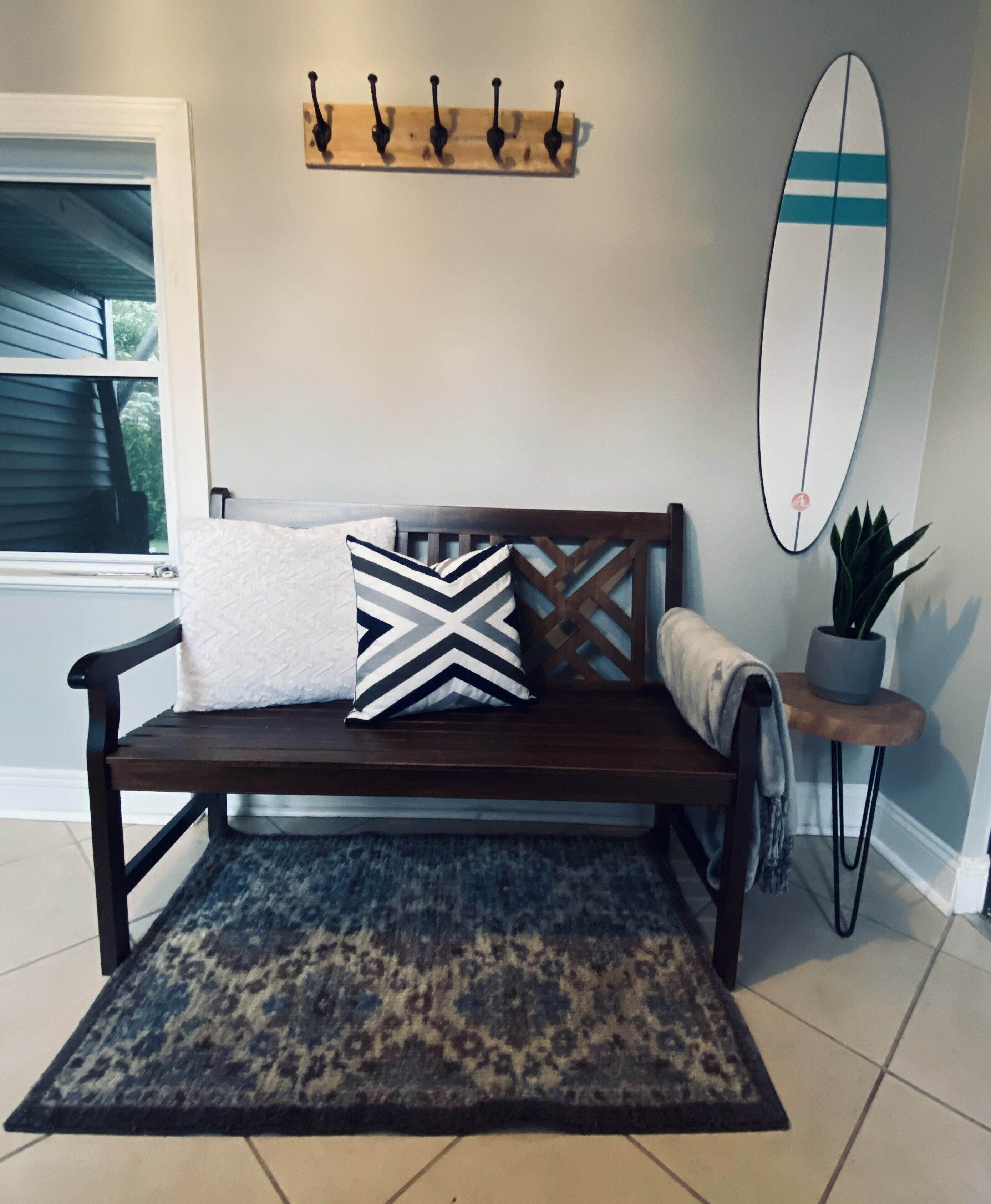 A wooden bench with a patterned rug, a wall-mounted surfboard, and a plant on a small table.