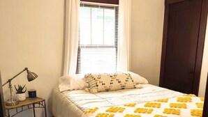 Un dormitorio ordenado con cama, mesita de noche con lámpara, ventana con cortinas y suelo de madera.