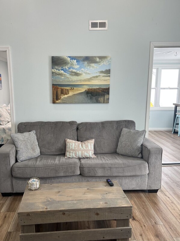 Un salon avec un canapé gris, une table basse en bois et un tableau sur le thème de la plage accroché au mur.