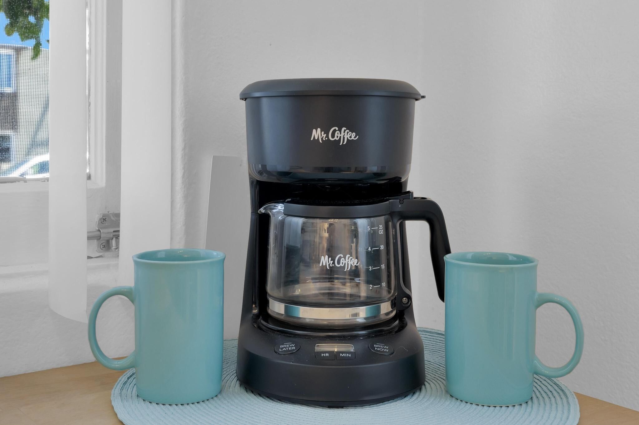 A black Mr. Coffee coffee maker with two turquoise mugs on a woven placemat.