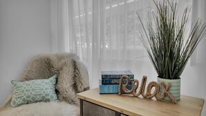 A cozy reading nook with a plush chair, a small table with books and a potted plant, and a window with sheer curtains. - #4286 PB Studio Walk to BAY, PIER & BEACH 🏝 (San Diego)
