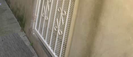 Una reja metálica blanca con diseños intrincados, una lámpara fijada a la pared y un muro de hormigón.