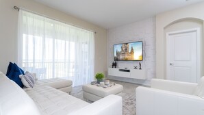 A modern living room with a white sofa set, a flat-screen TV mounted on a white brick wall, and a door to the right.