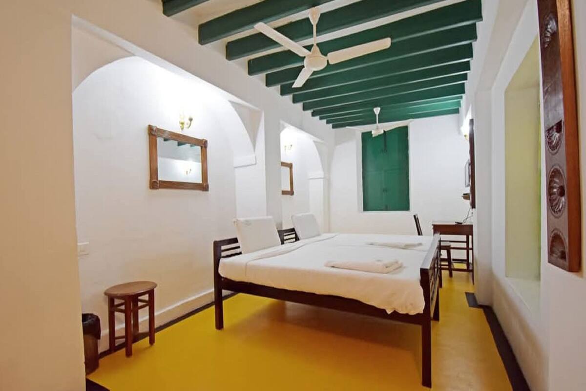 A bedroom with a bed, a small stool, a mirror, and a green window.