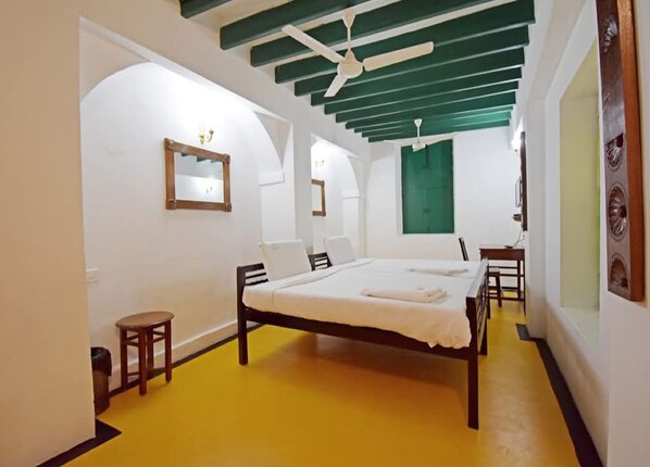 A bedroom with a bed, a small stool, a mirror, and a green window. - Villa Sentosa (Puducherry)