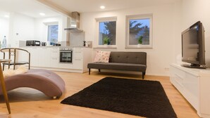 A modern kitchen with white cabinets, a grey sofa, a black rug, and a TV on a white cabinet.