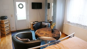 Une pièce comprenant un lit, une chaise, une petite table avec un vase, un téléviseur, une armoire et une fenêtre avec des rideaux.
