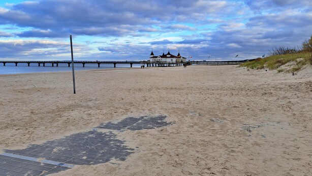 Ein Strand mit einem Steg, der ins Wasser führt, ein kleines Gebäude mit einem spitz zulaufenden Dach und ein Sandbereich mit einem Entwässerungsrost.