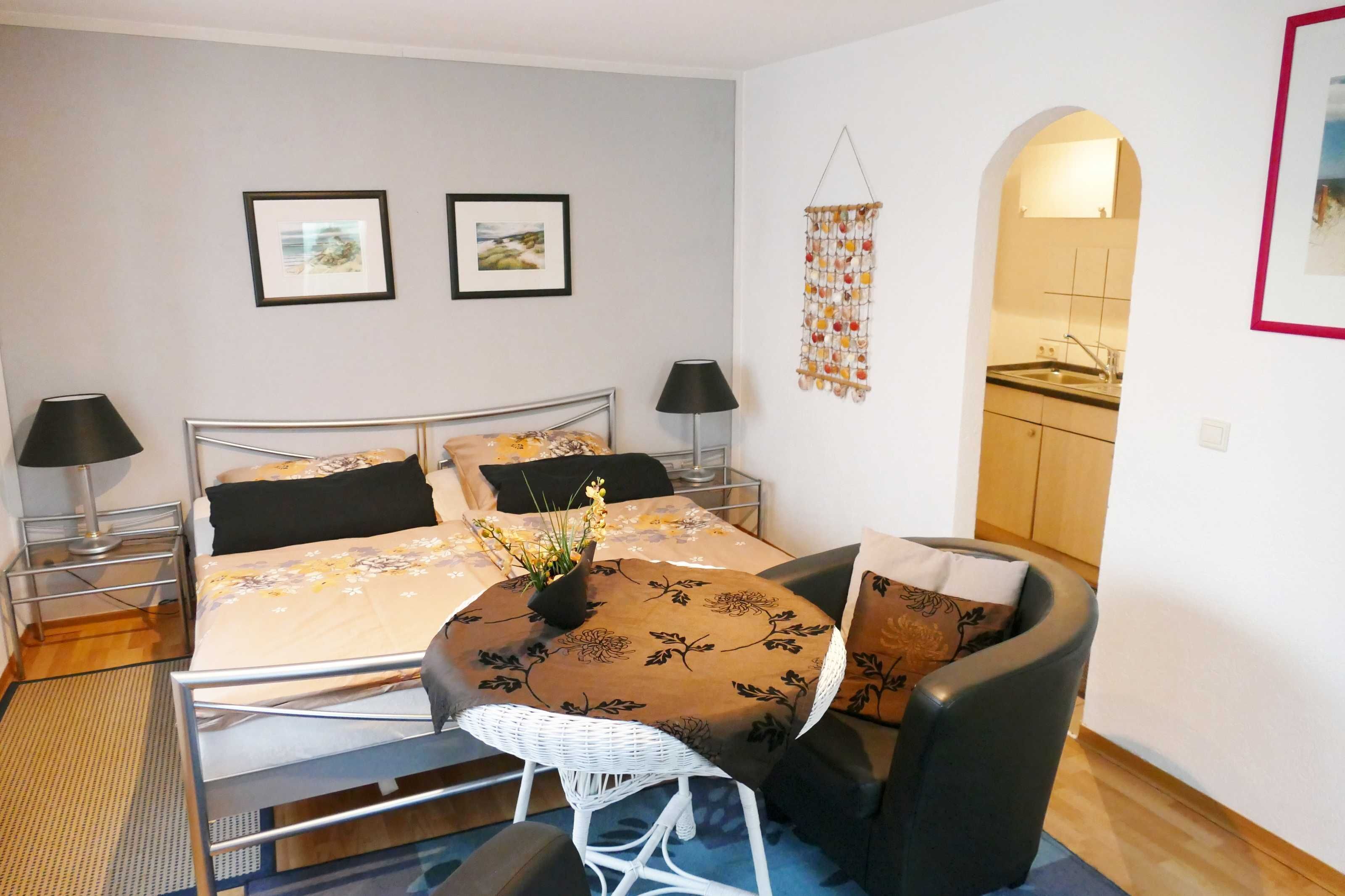 Une chambre à coucher de taille modeste, comprenant un lit, des tables de chevet, une petite table ronde avec un vase, une chaise et une vue sur la cuisine par le biais d’une arche.