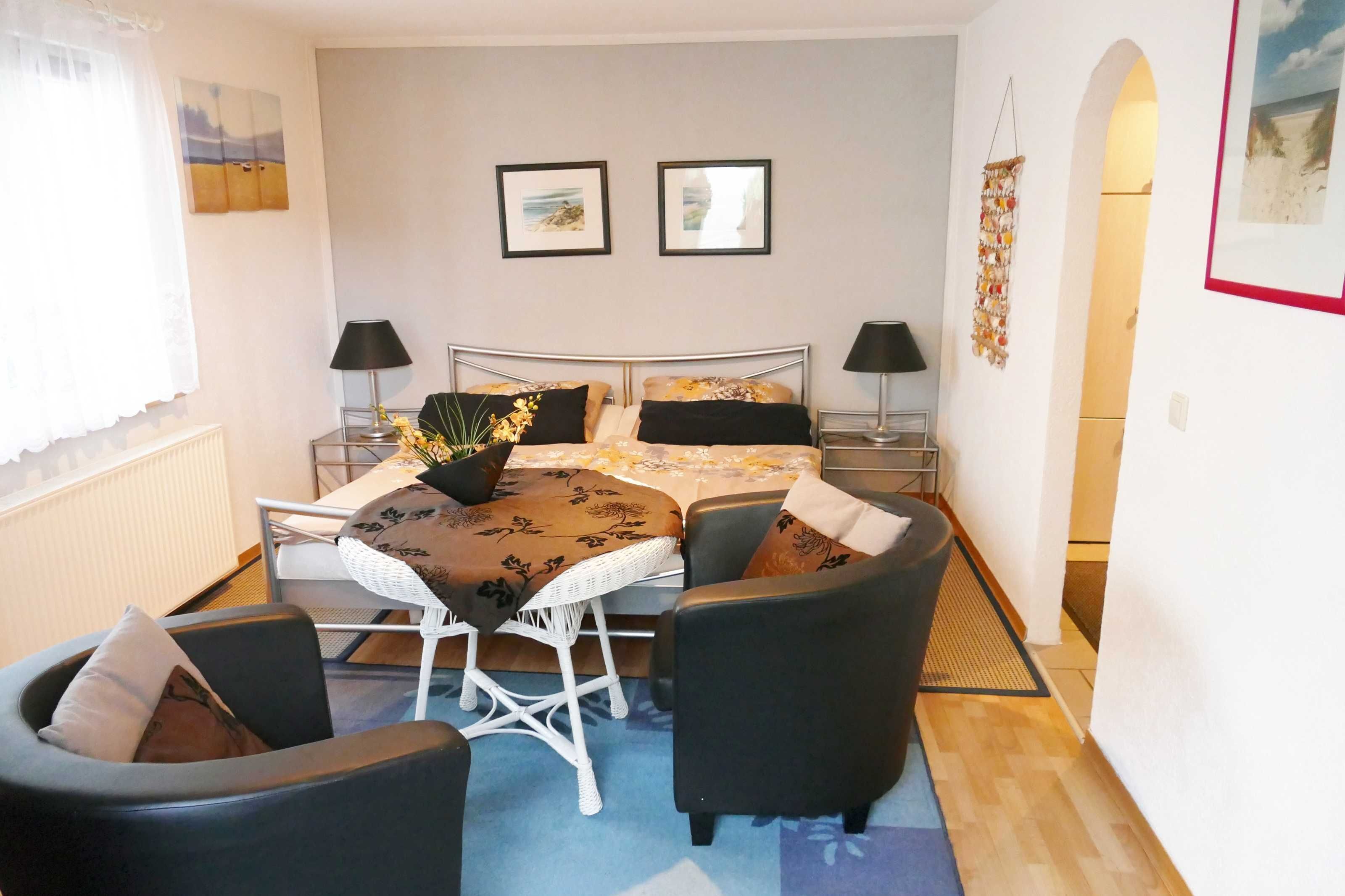 Ein Schlafzimmer mit einem Bett, zwei Nachttischen, einem Sessel, einem kleinen Tisch und einem Fenster mit Vorhängen.