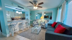 A living room with a grey sofa, red cushions, and patterned chairs. There is a kitchen area with white cabinets and a bar counter. - Fabulous 1BR Condo with amazing beach views sleeps 4. Park by door. Pool. (Panama City Beach)