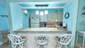 A kitchen with a bar area, featuring a refrigerator, a microwave, and a coffee maker. There are three white bar stools and a clock on the wall. - Fabulous 1BR Condo with amazing beach views sleeps 4. Park by door. Pool. (Panama City Beach)