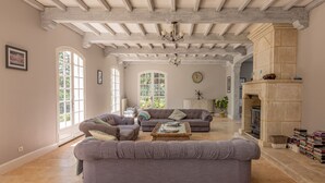 A spacious living room with a high ceiling, exposed beams, a fireplace, and large windows. - Quiet 50's cottage - 12 p. - Private park & pool (Fourcès)