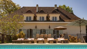 A two-story house with a swimming pool in front. There are lounge chairs and a table with umbrellas. - Quiet 50's cottage - 12 p. - Private park & pool (Fourcès)