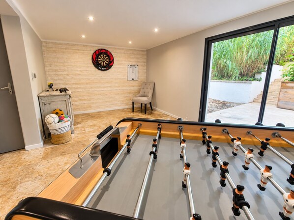 A foosball table in a room with a dartboard, a chair, and a basket with balls.