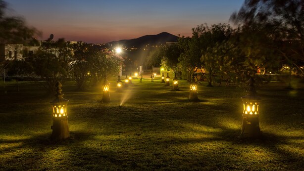 Escena nocturna al aire libre con una fila de faroles iluminados, un área con césped y luces de la ciudad a lo lejos.