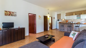 Una sala de estar moderna con un sofá, una mesa de centro y un televisor. Se ve una cocina al fondo, con taburetes de bar y armarios.