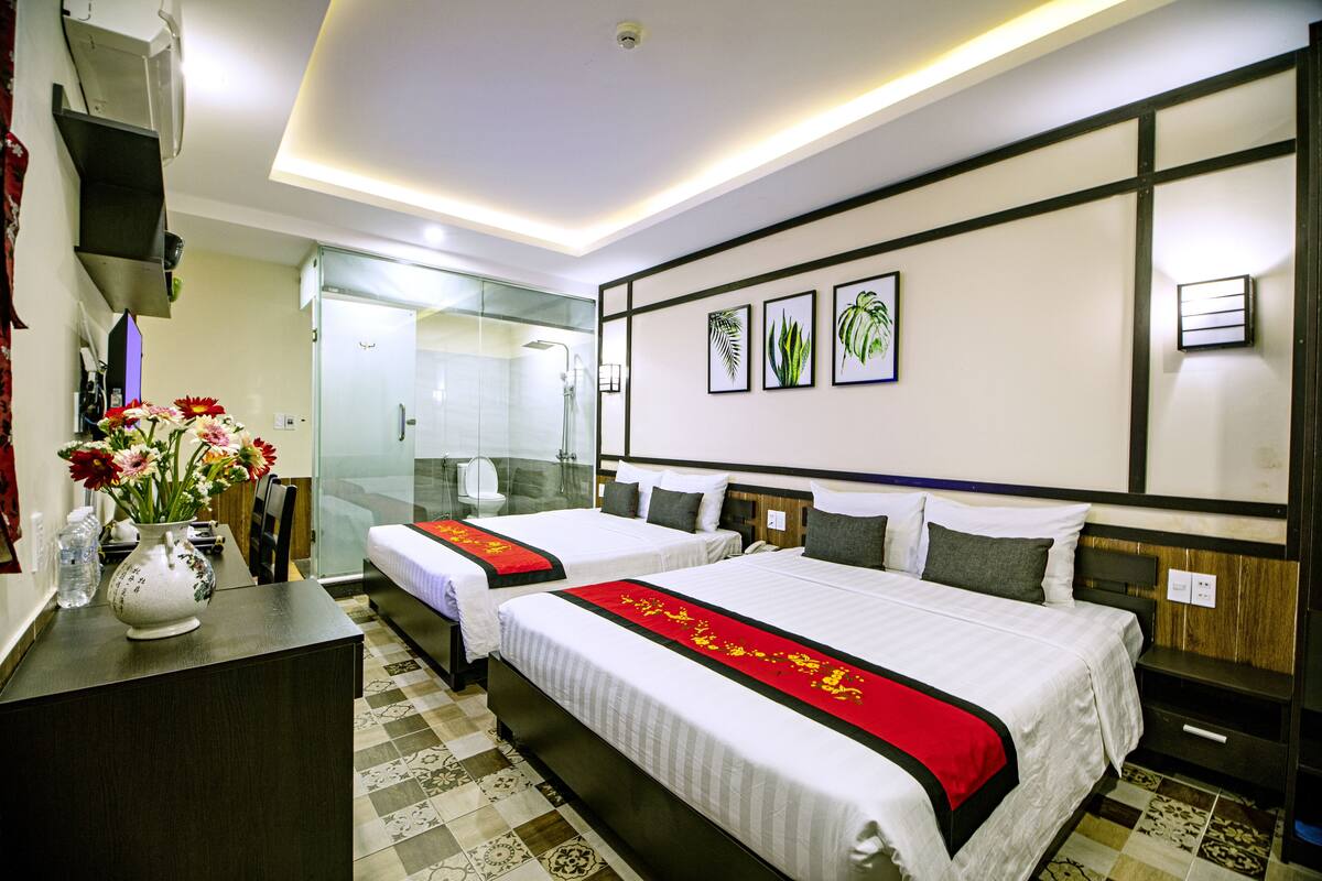 A hotel room with two beds, a shower, a desk with a vase of flowers, and framed artwork on the wall.