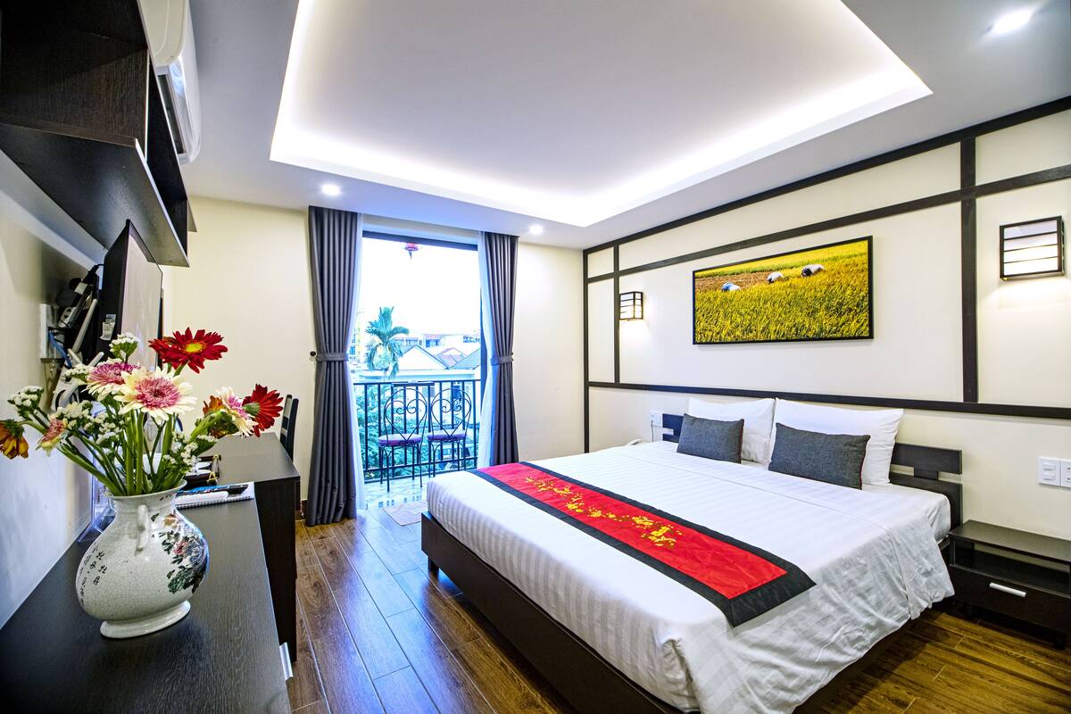 A hotel room with a large bed, a vase of flowers, a painting on the wall, and a balcony with a view.