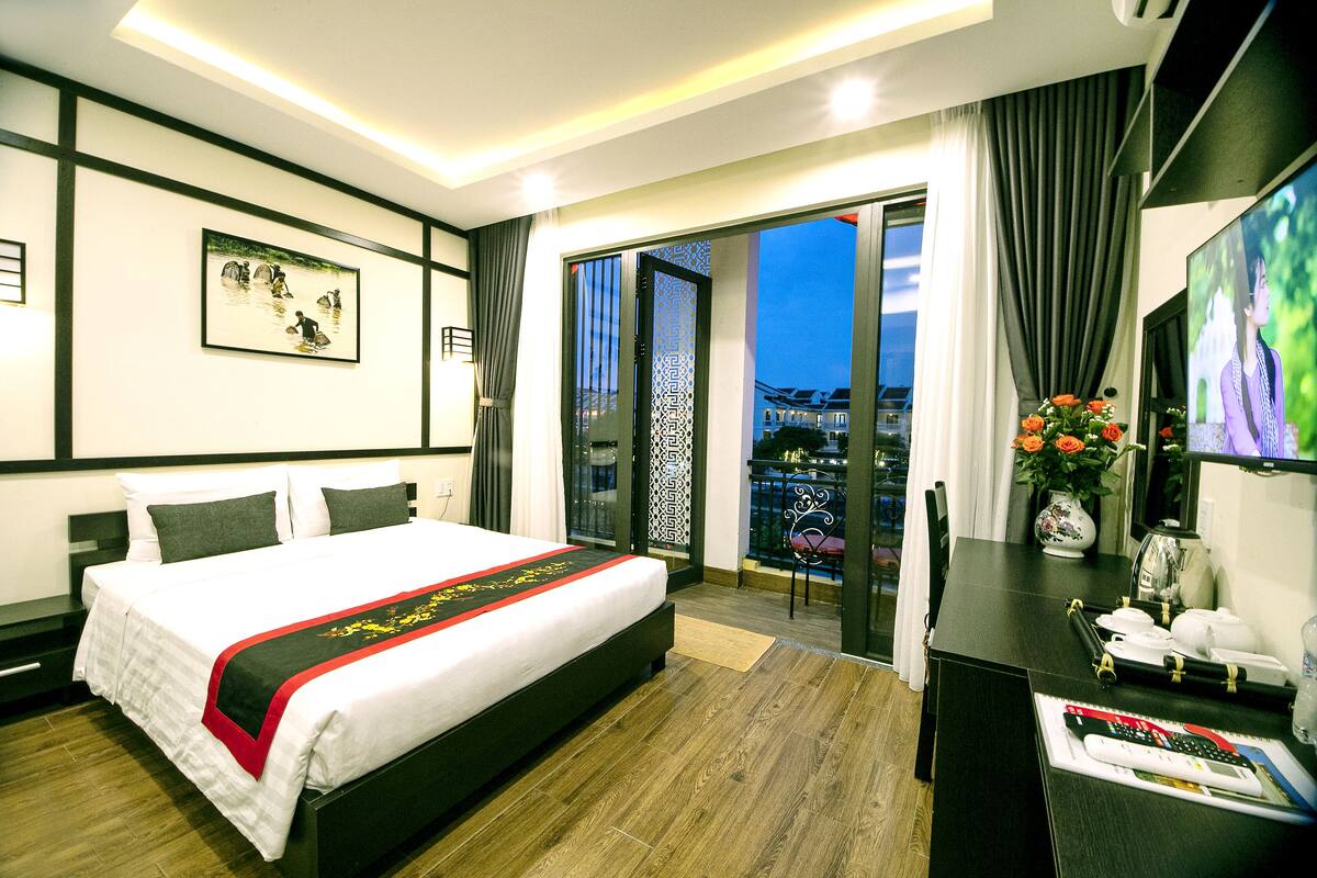 A hotel room with a large bed, a desk with a computer, a TV, and a balcony with a view.