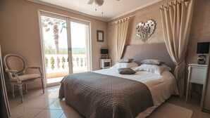 Un dormitorio con una cama grande, una silla, una mesita de noche, una lámpara y vistas a palmeras a través de un ventanal corredero.