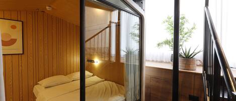 A bedroom with a bed, wooden walls, a window with curtains, a small table with a plant, and a staircase.