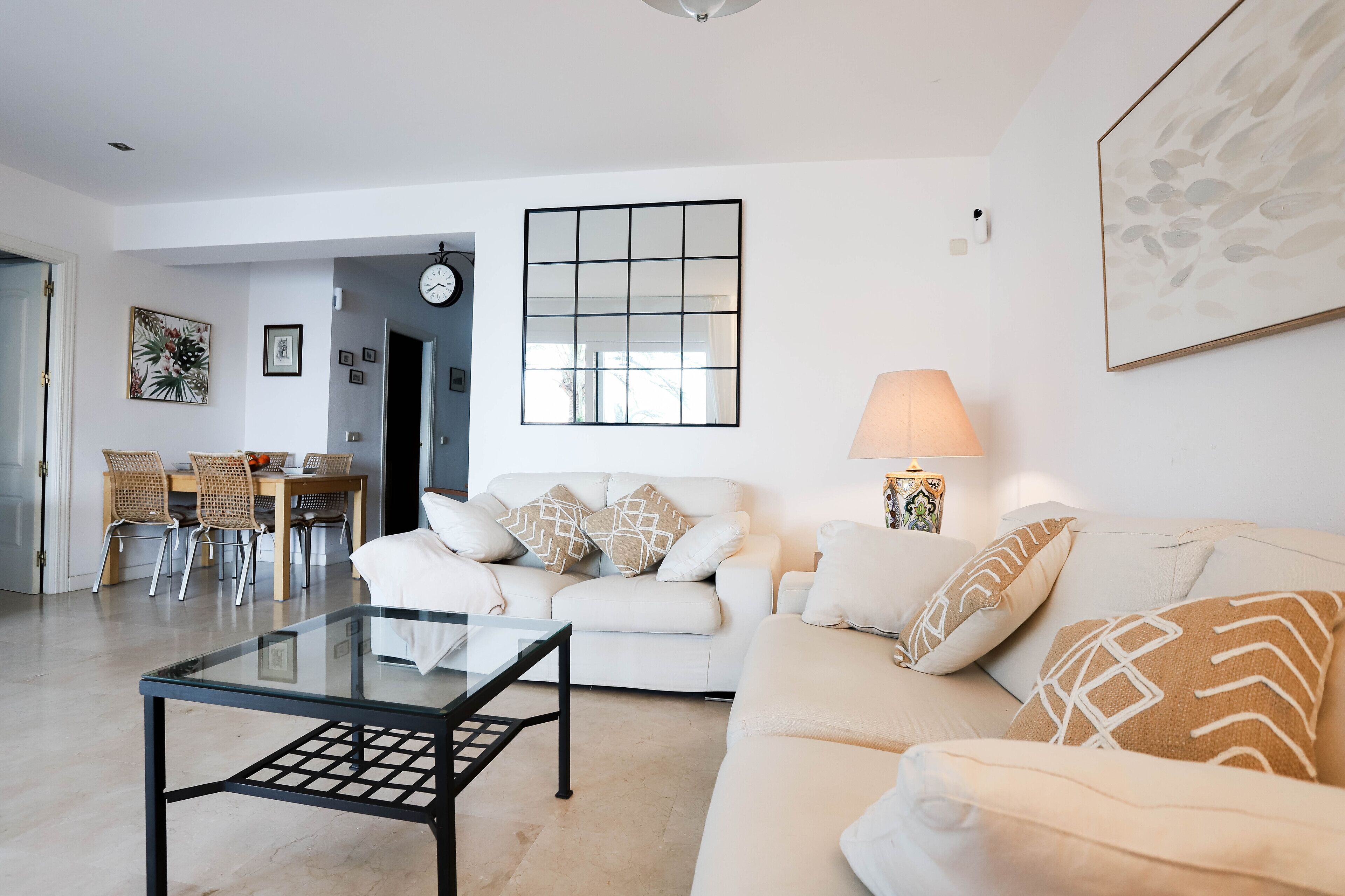 Een moderne woonkamer met een glazen salontafel, een witte bank en een eethoek met rieten stoelen.