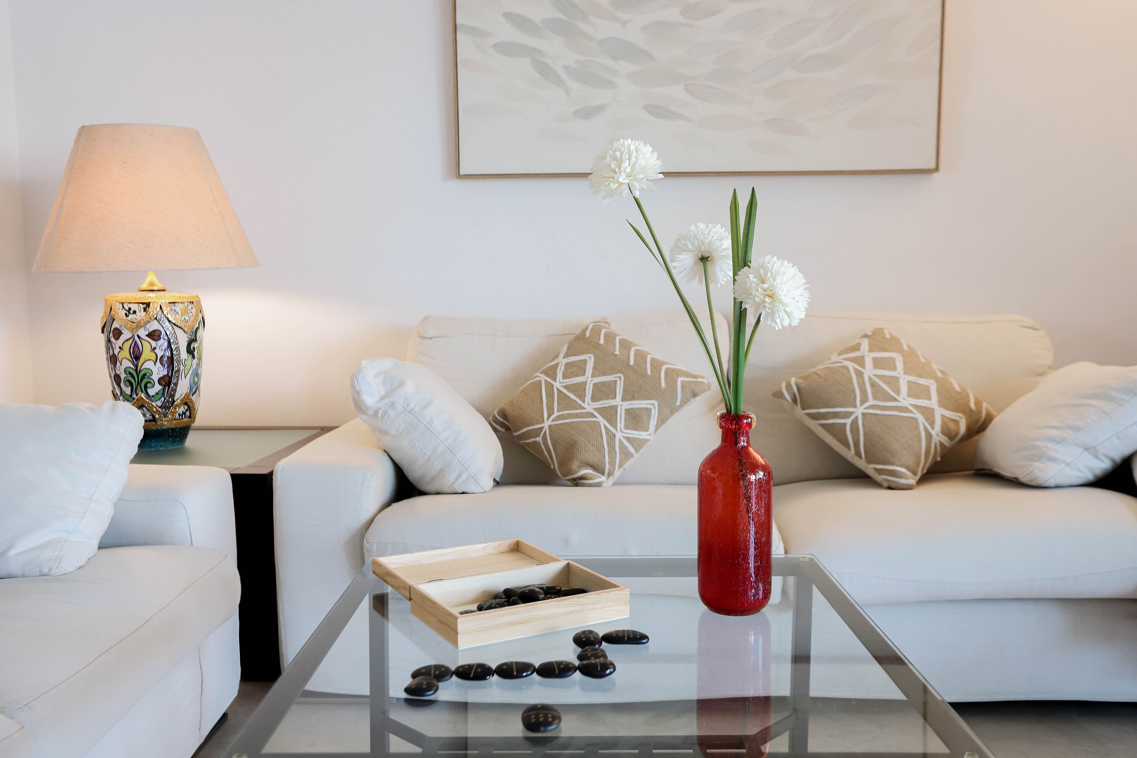 Ein modernes Wohnzimmer mit einem Glastisch, einer roten Vase mit weißen Blumen, einer dekorativen Lampe und einem geometrisch gemusterten Kissen auf einem weißen Sofa.