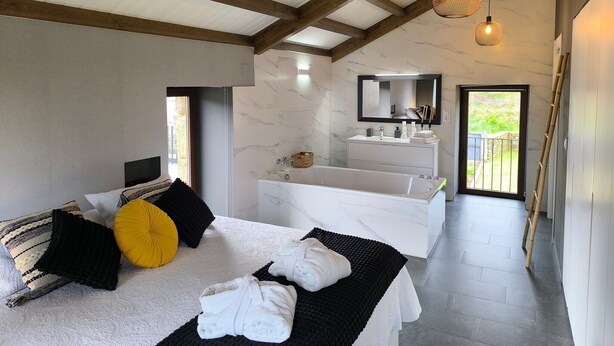 A modern bedroom with a large bed, a bathroom with a bathtub, and a view of the outdoors. - A Casa de Costa (Laxe)