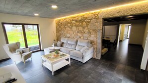 A modern living room with a stone accent wall, a grey sofa, a white coffee table, and a large window offering a view of a green outdoor area. - A Casa de Costa (Laxe)