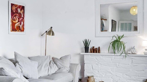 A living room with a white brick fireplace, a grey sofa, and a framed abstract painting. - Peaceful Home in Looe with Stunning Views (Looe)