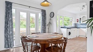 A dining area with a round wooden table and chairs, a hanging pendant light, and a view of the garden through large windows. - Peaceful Home in Looe with Stunning Views (Looe)