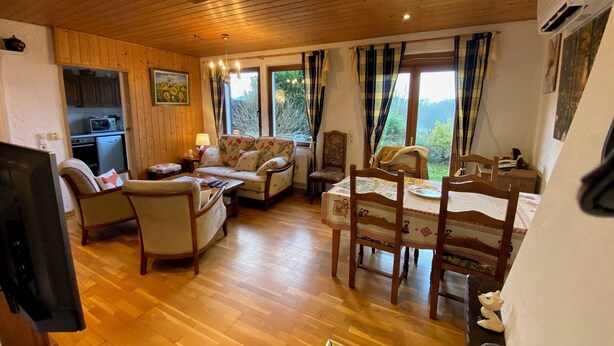 A cozy living room with wooden floors, a sofa, a dining table with chairs, and a kitchen area visible through an open door. - Gîte LE Petit Renard (Dabo)