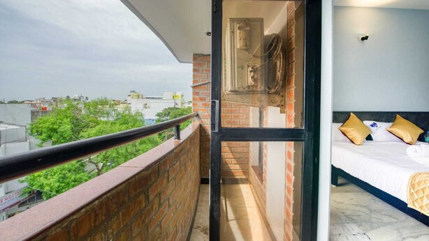 A balcony with a view of the city, a bed with a headboard, and a window with a view of a brick wall and electrical box. - Corbelli Residency  (Puducherry)
