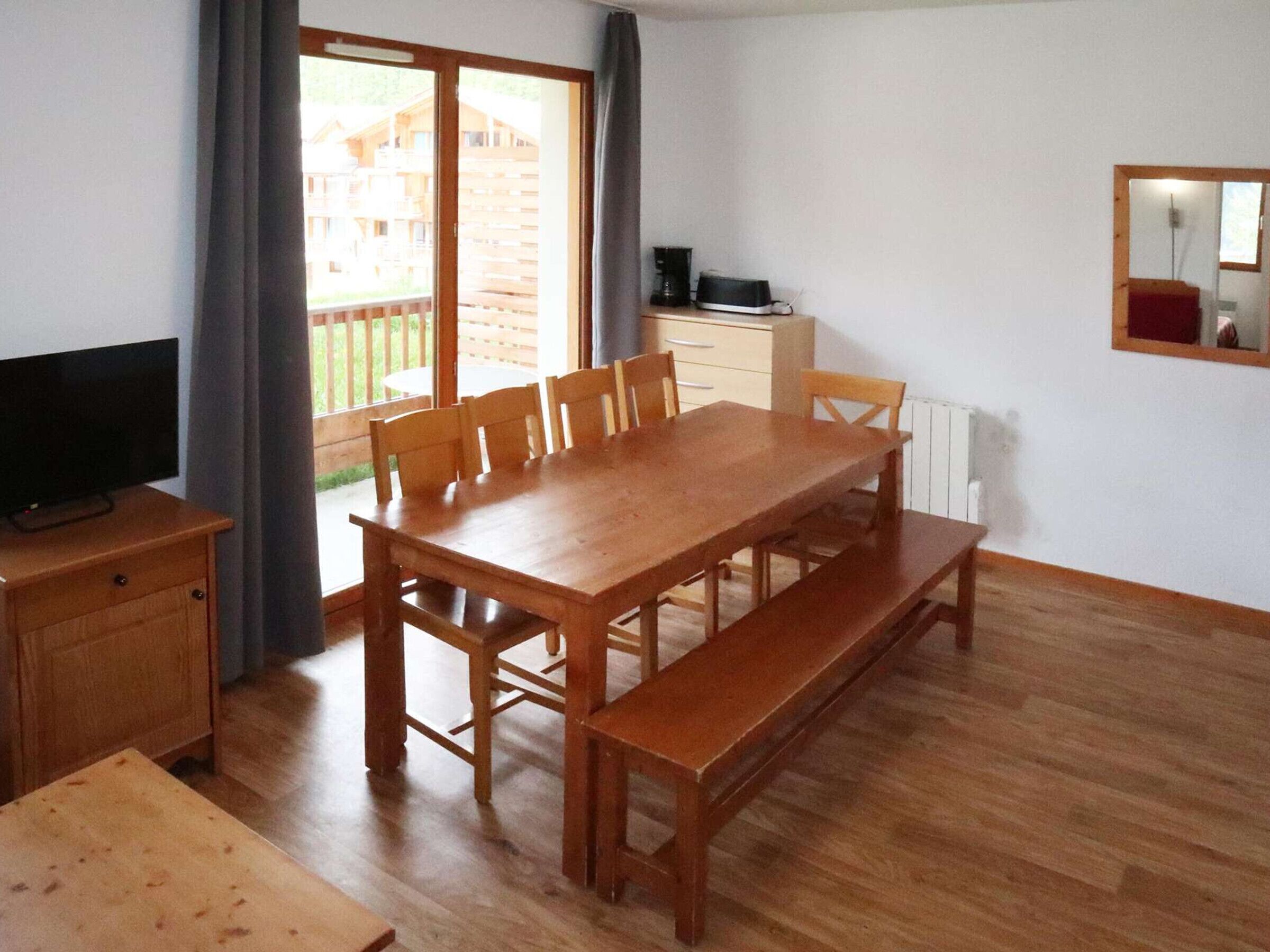 Un comedor con una mesa de madera y bancos, un televisor sobre un mueble bajo, y vista a una terraza de madera.