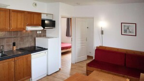 A compact kitchen with wooden cabinets, a microwave, a stove, and a dishwasher. There is a red sofa and a wooden table in the living area. - Les Orres 1800: 3-room Apt, Pool, WiFi, Pets allowed (Les Orres)
