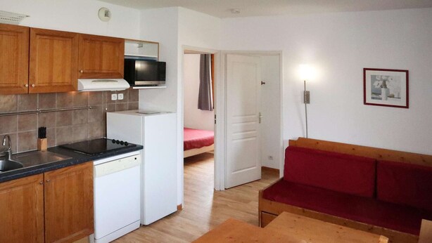 A compact kitchen with wooden cabinets, a microwave, a stove, and a dishwasher. There is a red sofa and a wooden table in the living area.