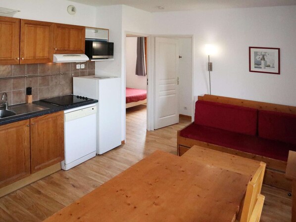 Cocina compacta con armarios de madera, microondas, estufa y lavavajillas. En la sala de estar, hay un sofá rojo y una mesa de madera.