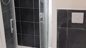 A glass-enclosed shower with a handheld showerhead, black tiled walls, and a white light switch on the wall. - Budget Ensuite Room in Thamesmead (London)