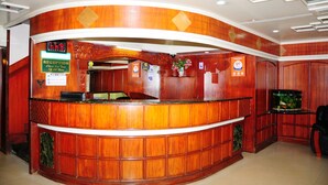 A reception area with a wooden reception desk, a digital clock, and a sign indicating check-out time. - Hotel Venus (Ootacamund)