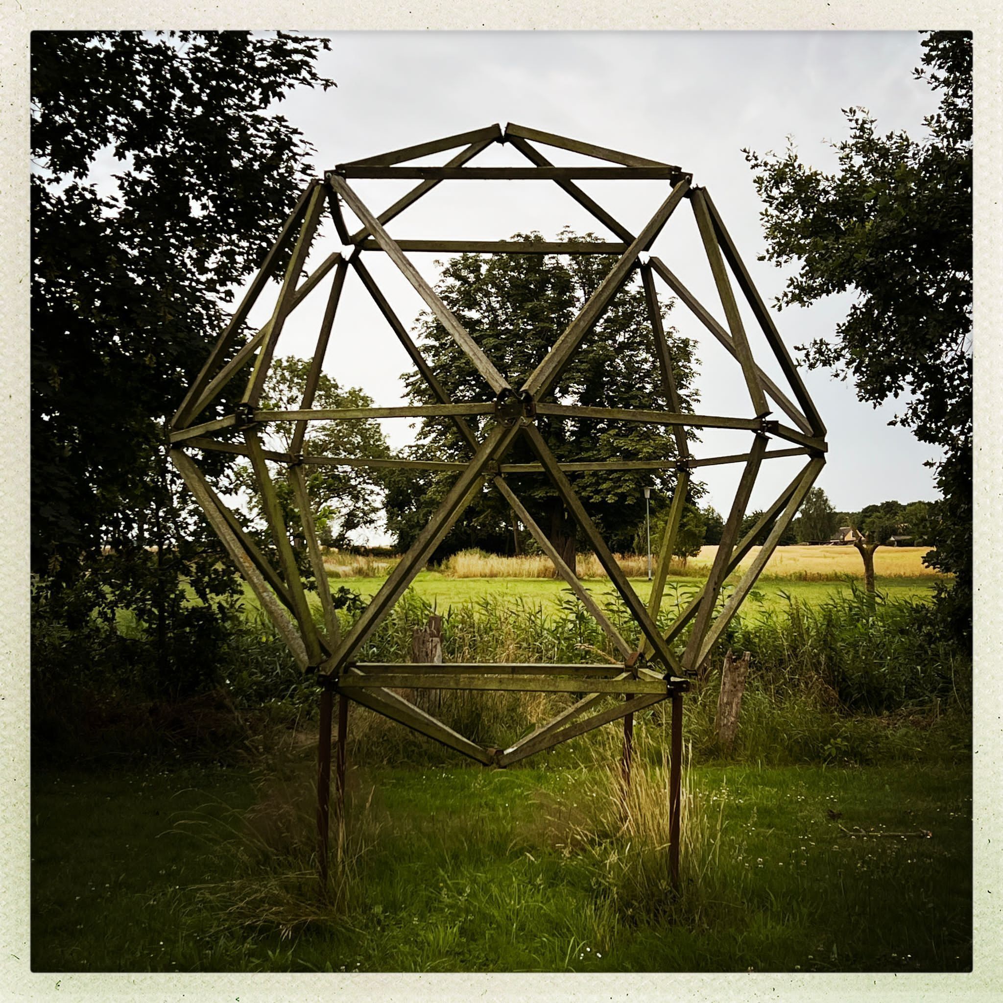 A geometric metal structure in a grassy field with trees in the background.