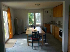 Een kleine keuken met een eettafel, stoelen, een koelkast en een raam met gordijnen.