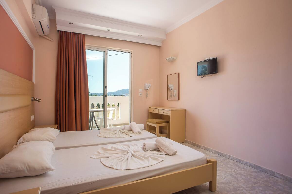 A hotel room with two beds, a desk, a chair, a TV, and a view of the outdoors.