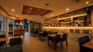 A restaurant with tables and chairs, a bar area with shelves of bottles, and a view through the glass doors. - Diagoras Hotel (Rhodes)