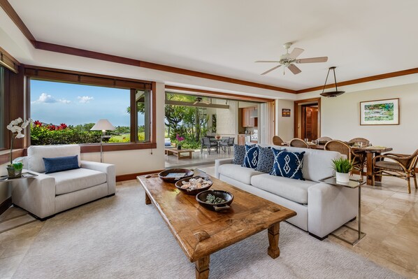 A spacious living area with a large window, a wooden coffee table, two sofas, and a dining area with wicker furniture.