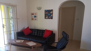 Un salon avec un canapé en cuir noir, deux coussins rouges, une table basse en bois et un fauteuil de bureau noir. Deux œuvres d’art ornent le mur, l’une représentant des poissons et l’autre un crabe. On aperçoit une porte au fond.