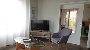 A modern living room with a glass-top coffee table, a grey armchair, a flat-screen TV on a wooden stand, and a view of the kitchen through a glass door. - Landliebeehrsen 2, modern, quiet location, parking lot.  (Bad Salzuflen)